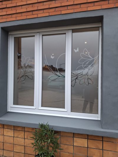 Fenêtre blanche à trois vantaux d'une façade en briques rouges dont le bas du vitrage porte un film à effet sablé ajouré de grandes corolles, de tiges courbes et de papillons