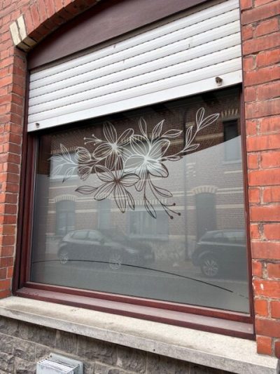 Fenêtre d'une façade en briques rouges, sous un volet roulant, dont le vitrage porte un film à effet sablé dessinant de grandes fleurs au trait, des feuilles et des boutons