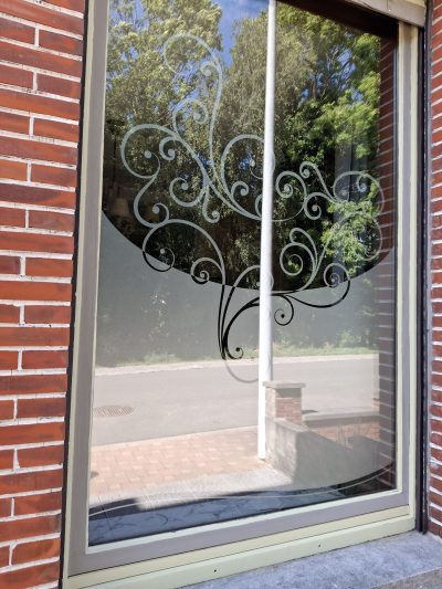 Grande fenêtre fixe d'une façade en briques rouges dont la partie basse porte un film à effet sablé au bord bombé, ajouré d'un arbre en volutes dont les branches se prolongent en spirales sablées sur le verre clair