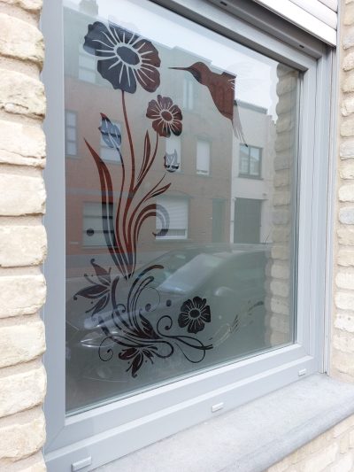 Fenêtre à châssis gris d'une façade en briques claires avec un film à effet sablé ajouré de grandes fleurs sur tige, d'un colibri et de volutes