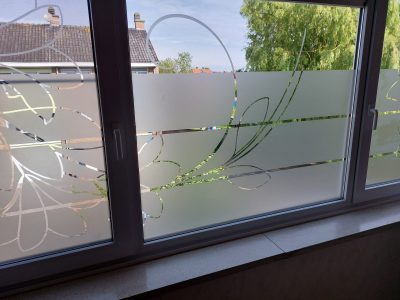 Fenêtre blanche à trois vantaux vue de l'intérieur, dont le vitrage porte un film à effet sablé ajouré d'une grande fleur stylisée, de longues feuilles arquées et d'un cercle