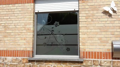 Fenêtre d'une façade en briques claires avec un film à effet sablé ajouré d'une tige fleurie en volutes et de fines bandes horizontales