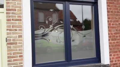 Fenêtre à deux vantaux à châssis bleu marine dans une façade en briques, dont le vitrage porte un film à effet sablé à bord ondulé ajouré de rinceaux, de feuilles et de marguerites
