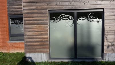 Baie vitrée à deux vantaux et petite fenêtre d'une façade en bardage bois et briques, avec un film à effet sablé bordé de volutes et de feuilles