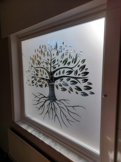 Fenêtre blanche vue de l'intérieur dont le vitrage porte un film à effet sablé ajouré d'un arbre de vie aux feuilles et aux racines déployées