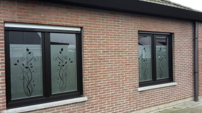 Deux fenêtres à châssis noirs d'une façade en briques rouges dont les quatre vantaux portent un film à effet sablé à motif de tiges feuillues et de volutes