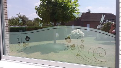 Fenêtre donnant sur un jardin, dont le vitrage porte un film à effet sablé en bandeau ondulé ajouré de fleurs et de volutes, surmonté de papillons et d'une libellule