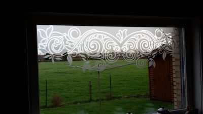 Grande fenêtre vue de l'intérieur, donnant sur un jardin et un champ, dont le haut du vitrage porte un film à effet sablé ajouré de volutes, de feuillages et de fleurs