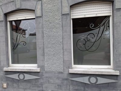 Deux fenêtres à volets roulants d'une façade grise crépie à encadrements moulurés dont le vitrage porte un film à effet sablé en partie basse, prolongé de volutes et de pois sablés et rehaussé d'arabesques noires