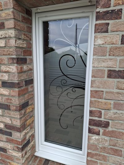 Fenêtre étroite à châssis blanc dans une façade en briques rouges, dont le vitrage porte un film à effet sablé ajouré de grandes volutes et d'arabesques