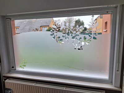 Fenêtre d'une pièce vue de l'intérieur, au-dessus d'un radiateur, avec un film à effet sablé à bord ondulé ajouré de feuillage, de vrilles et d'une jeune pousse