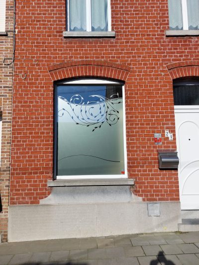Fenêtre sous arc de briques d'une façade en briques rouges dont le vitrage porte un film à effet sablé bordé de rinceaux feuillus ajourés et traversé d'une courbe