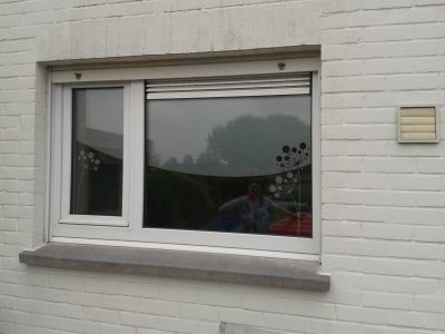 Fenêtre à deux vantaux d'une façade en briques peintes en blanc dont les vitrages portent trois aigrettes de pissenlit découpées dans un film à effet sablé