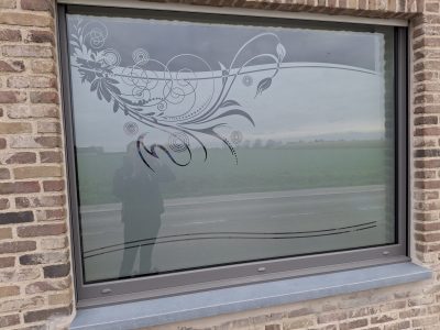Grande fenêtre à châssis gris d'une façade en briques avec un film à effet sablé à motif de volutes fleuries, de cercles concentriques et de lignes ondulées