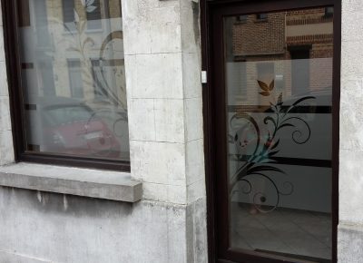 Porte d'entrée en bois et fenêtre d'une façade en pierre claire, la porte avec une bande à effet sablé à mi-hauteur et les deux vitrages avec un motif de volutes et de feuilles en film foncé