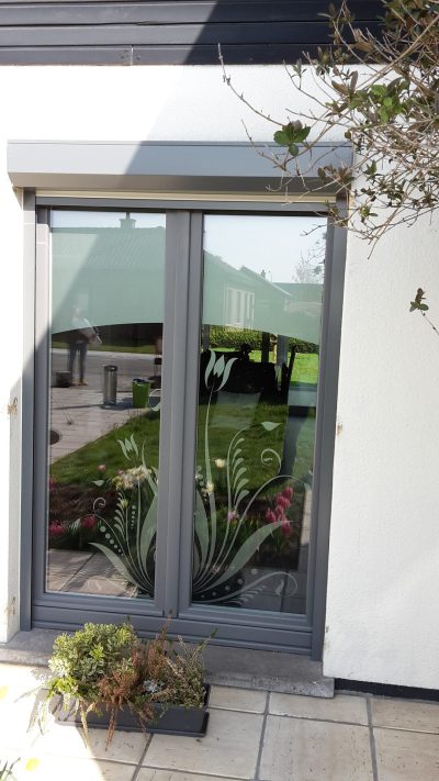 Grande fenêtre à deux vantaux au châssis gris anthracite, sur une façade blanche, dont le bas du vitrage porte un film à effet sablé ajouré d'une gerbe de fleurs, de longues feuilles et de volutes