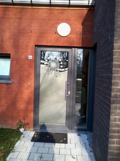 Porte d'entrée en aluminium anthracite d'une maison en briques rouges dont le vitrage porte un film à effet sablé ajouré d'un pissenlit et de courbes ondulantes