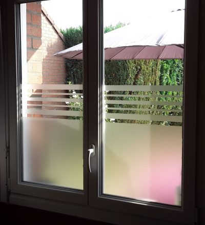 Fenêtre à deux vantaux vue de l'intérieur, film à effet sablé surmonté de bandes horizontales, jardin visible au-dessus