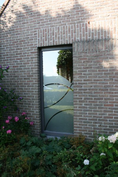Porte vitrée d'une façade en briques claires, film à effet sablé à motif d'arcs de cercle, massif fleuri devant