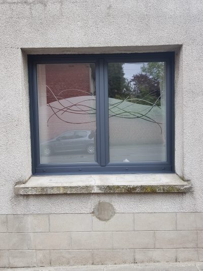 Fenêtre à deux vantaux gris anthracite d'une façade crépie blanche, dont le vitrage porte un film à effet sablé à bord supérieur arqué, traversé d'un faisceau de courbes entrelacées