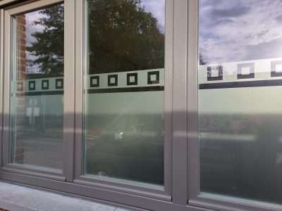 Fenêtre à châssis gris anthracite d'une façade en briques, dont la moitié inférieure des vitrages porte un film à effet sablé bordé d'une frise de petits carrés ajourés