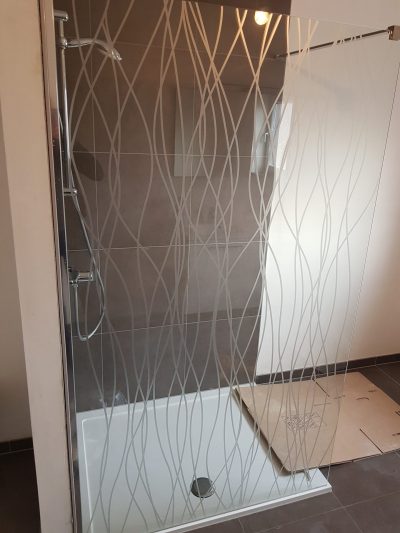 Paroi de douche vitrée d'une salle de bain carrelée de gris avec un film à effet sablé à motif de lignes ondulées entrelacées