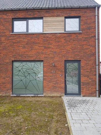 Façade en briques rouges d'une maison dont la baie vitrée, la porte d'entrée et les fenêtres de l'étage portent un film à effet sablé ajouré de lignes brisées formant des facettes triangulaires