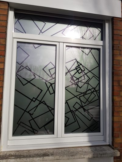Fenêtre blanche à deux vantaux et imposte sur une façade en briques rouges, avec un film à effet sablé à motif de rectangles entrecroisés tracés en noir