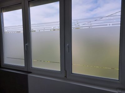Fenêtre blanche à trois vitrages vue de l'intérieur, dont le film à effet sablé s'arrête sous une frise de traits horizontaux décalés et laisse un fin filet transparent vers le bas