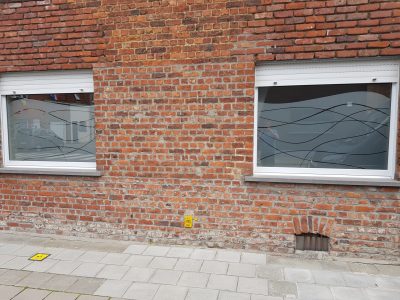 Deux fenêtres à volets roulants d'une façade en briques rouges dont le vitrage porte un film à effet sablé au bord supérieur ondulé, traversé de lignes courbes