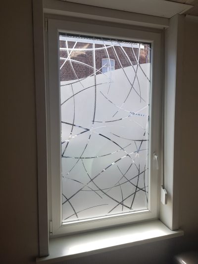 Fenêtre en PVC blanc vue de l'intérieur dont le vitrage porte un film à effet sablé au bord supérieur en vague, ajouré de fins arcs entrecroisés qui se prolongent en arcs sablés sur la partie claire