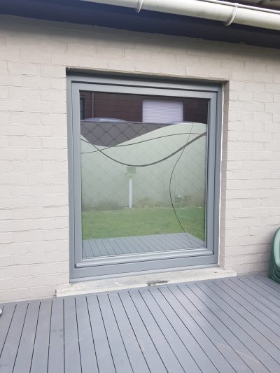 Grande fenêtre à châssis gris sur un mur de briques peintes en blanc, dont le vitrage porte un film à effet sablé au bord supérieur ondulé, traversé de deux fines courbes