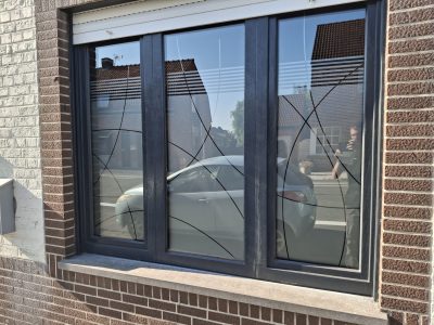 Fenêtre à trois vantaux gris anthracite d'une façade en briques brunes, dont le vitrage porte un film à motif de courbes entrecroisées sous une bande de fines rayures à effet sablé