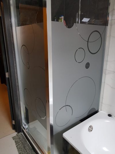 Cabine de douche d'angle à cadre chromé dont les deux vitrages portent un film à effet sablé ajouré de cercles et de disques de tailles variées, le haut du verre restant transparent
