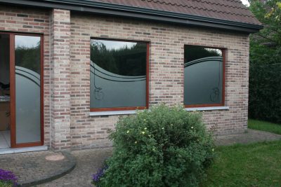 Porte vitrée et deux fenêtres à châssis bois d'une façade en briques claires dont le bas du vitrage porte un film à effet sablé à bord en vague, ajouré d'un trait fin et d'une volute
