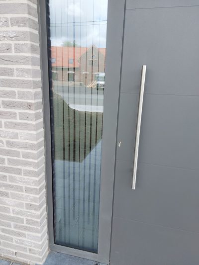 Vitrage latéral d'une porte d'entrée gris anthracite, sur une façade en briques grises, avec un film à effet sablé à fines lignes verticales