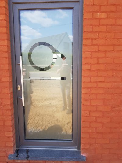 Porte d'entrée grise dans une façade en briques rouges avec un film à effet sablé ajouré d'un grand anneau prolongé d'un trait brisé et d'une bande horizontale
