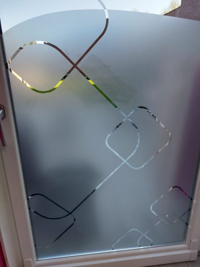 Ouvrant cintré à cadre blanc, vu de l'intérieur, dont le vitrage porte un film à effet sablé ajouré de losanges arrondis, d'une boucle croisée et d'une longue diagonale