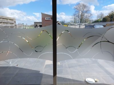 Grande baie vitrée à deux vantaux donnant sur une terrasse dallée, dont le vitrage porte un bandeau de film à effet sablé aux bords ondulés, ajouré de rubans courbes qui se croisent en vagues