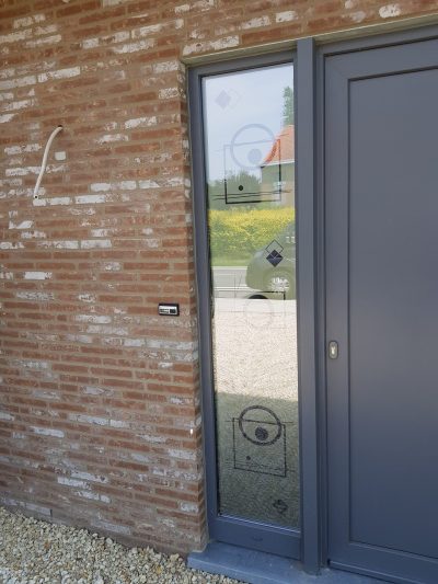 Fixe latéral d'une porte d'entrée grise sur une façade en briques rouges, dont le vitrage porte un film à effet sablé à motifs géométriques de losanges, de cercles et de cadres