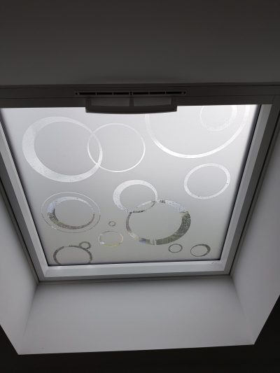 Fenêtre de toit vue de l'intérieur dont le vitrage porte un film à effet sablé ajouré d'anneaux de tailles variées