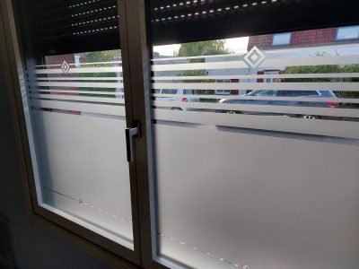 Fenêtre à deux vantaux vue de l'intérieur, dont le vitrage porte un film à effet sablé plein en partie basse, terminé par des bandes horizontales de plus en plus fines et un losange dans l'angle supérieur gauche de chaque vantail