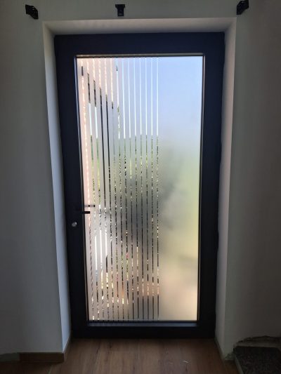 Porte d'entrée vitrée à cadre anthracite, vue de l'intérieur, dont le vitrage porte un film à effet sablé ajouré de fines rayures verticales qui s'espacent vers la droite