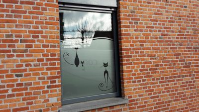 Fenêtre à châssis gris d'une façade en briques rouges avec un film à effet sablé au bord supérieur découpé en arc, orné de trois silhouettes de chats noirs à la queue en volute