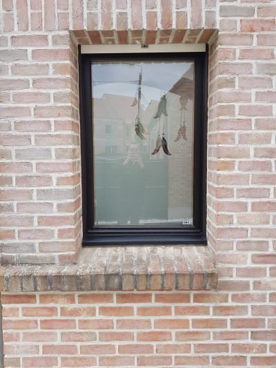 Fenêtre à châssis noir d'une façade en briques rose pâle dont le vitrage porte un film à effet sablé au bord supérieur arqué, ajouré de plumes suspendues à des fils