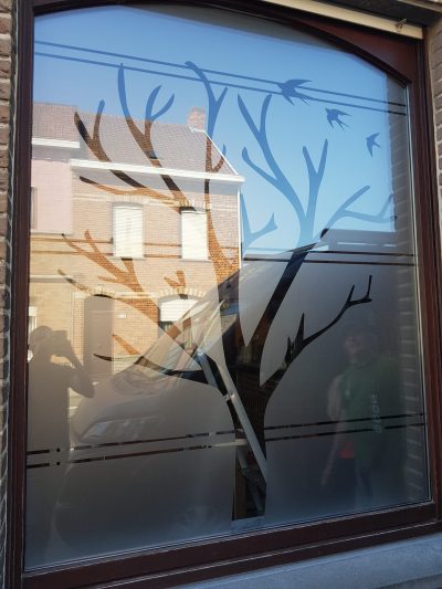 Grande fenêtre cintrée d'une maison en briques avec un film à effet sablé à motif d'arbre aux branches nues et d'hirondelles, le tronc découpé dans une bande pleine