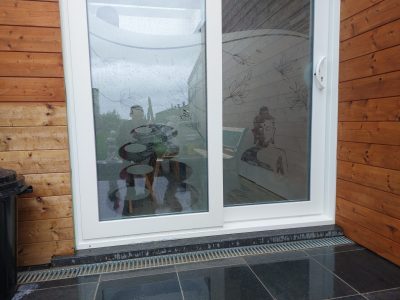 Baie coulissante blanche à deux vantaux dans une façade en bardage bois, dont le vitrage porte un film à effet sablé à motif de Bouddha et de fleurs