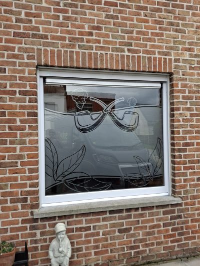 Fenêtre d'une façade en briques rouges dont le vitrage porte un film à effet sablé dessinant deux pipes croisées fumantes et de grandes feuilles nervurées