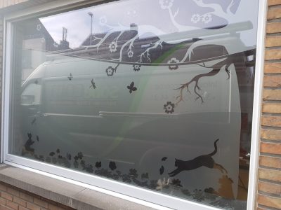 Grande fenêtre d'une façade en briques avec un film à effet sablé ajouré de chats et de papillons, surmonté de branches de cerisier en fleurs et d'une chouette