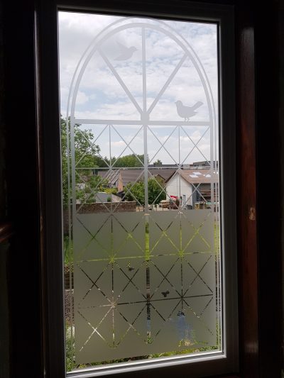 Fenêtre à cadre blanc vue de l'intérieur avec un film à effet sablé dessinant une arche treillagée, deux oiseaux et quelques papillons, la partie basse en aplat plein